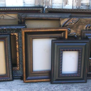 May include: A collection of multiple rectangular picture frames in various sizes and finishes, including black, dark brown, and metallic bronze tones. The frames feature ornate, textured, and smooth borders, some with intricate embossed patterns.