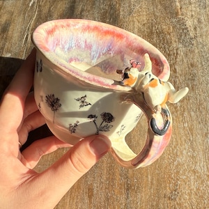 Peut inclure: Une tasse en céramique à base blanche, ornée d'illustrations florales et de papillons noirs. Le bord a une finition rose et irisée. Une petite figurine de chat est attachée à la poignée. La tasse est tenue par une main.