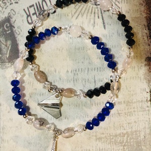 May include: Two beaded bracelets with a silver Eiffel Tower charm and a silver paper airplane charm. The bracelets are made with black, blue, and clear beads.