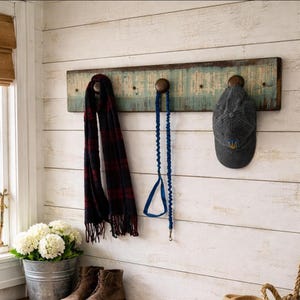 May include: A rustic wooden wall-mounted coat rack with three dark wooden pegs. A navy and burgundy plaid scarf, a blue dog leash, and a gray baseball cap with a yellow emblem hang from the pegs. The rack has a distressed blue and brown finish.