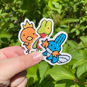 May include: Three cartoon stickers: a chick with a crown, a frog, and a blue creature with orange accents. The stickers are held against a backdrop of green leaves, showcasing the vibrant colours and playful designs.