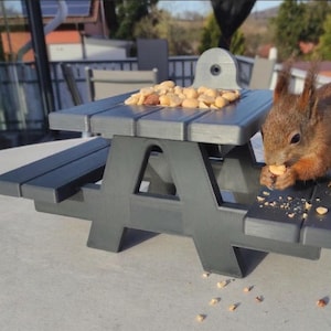 May include: A dark grey miniature picnic table feeder filled with peanuts. A red squirrel is eating a peanut on the table. The feeder is designed for outdoor use and is placed on a concrete surface.