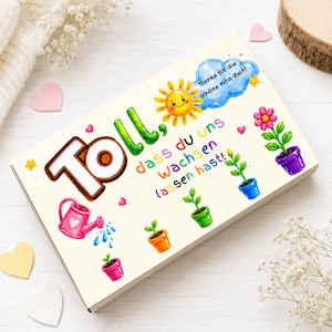 Pode incluir: Uma caixa retangular branca com o texto em alemão Toll, dass du uns wachsen lassen hast! e Danke für die schöne Kita-Zeit!, decorada com um sol amarelo, um regador rosa e várias plantas coloridas em vasos e pequenos corações.