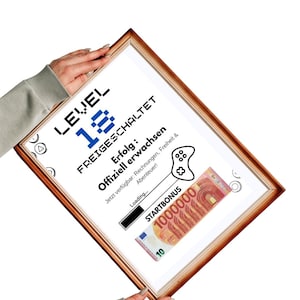 May include: A framed print with a brown wooden frame. The print features the text "LEVEL 18 FREIGESCHALTET" in black and blue pixelated font. Below, it reads "Erfolg: Offiziell erwachsen" and includes a graphic of a game controller and a 10 Euro note.