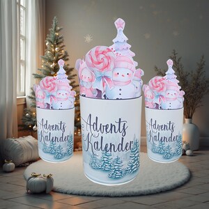 Könnte beinhalten: Drei weiße Adventskalender-Behälter mit winterlichen Illustrationen. Jeder Behälter zeigt eine Waldszene am Boden und die Worte "Advents Kalender" in einer Schreibschrift. Die Oberteile haben Ausschnitte von Schneemännern, Lollis und Weihnachtsbäumen.