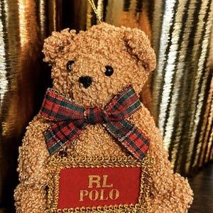 Puede incluir: Un adorno de oso de peluche texturizado de color canela con un lazo de cinta a cuadros rojos y verdes alrededor del cuello. El oso sostiene un letrero rectangular rojo con un borde trenzado dorado que muestra el texto dorado RL POLO.