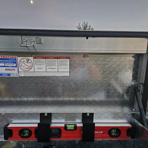 May include: A red and black level tool is mounted inside a metal truck bed storage compartment. The level has three bubble vials and is held in place by black brackets. Safety and warning labels are visible on the compartment wall.