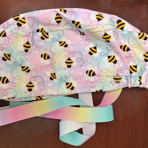 Op de afbeelding: Een pastelkleurige operatiemuts met een patroon van gele en zwarte bijen en witte bloemen. De muts heeft een bijpassend regenbooglint om te strikken. De stof is een mix van roze, blauw en geel.
