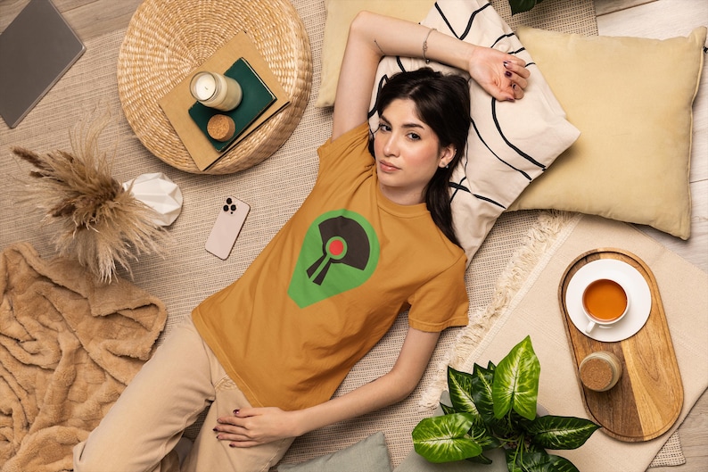 May include: Mustard yellow t-shirt with a green and black graphic design. The person is lying on a rug with pillows, a woven mat, a candle, and a smartphone. A cup of tea sits on a wooden tray.