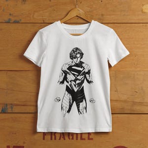 Puede incluir: Camiseta blanca con una ilustración en blanco y negro de un superhéroe, que muestra el emblema "S". El diseño muestra una figura musculosa en una pose dinámica. La camiseta está colgada en una percha de madera.