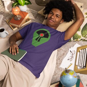 May include: A purple t-shirt featuring a green and black graphic with a red circle. The person is lying down on a rug with a globe, books, and a typewriter. The shirt is short-sleeved.