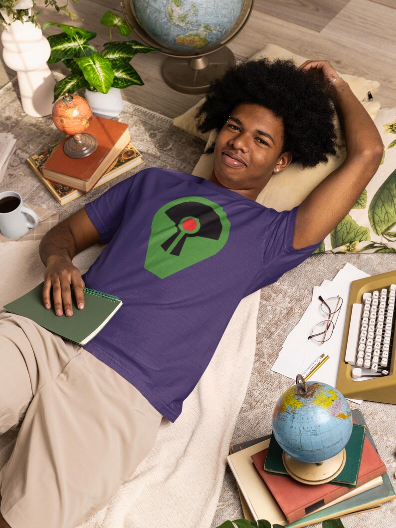 May include: A purple t-shirt featuring a green and black graphic with a red circle. The person is lying down on a rug with a globe, books, and a typewriter. The shirt is short-sleeved.