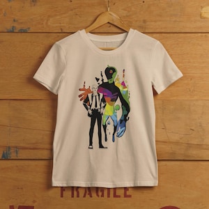 Puede incluir: Una camiseta color crema con un diseño gráfico de dos figuras. Una figura es un hombre con traje, y la otra es una forma abstracta colorida. El diseño incorpora un vibrante arcoíris de colores.