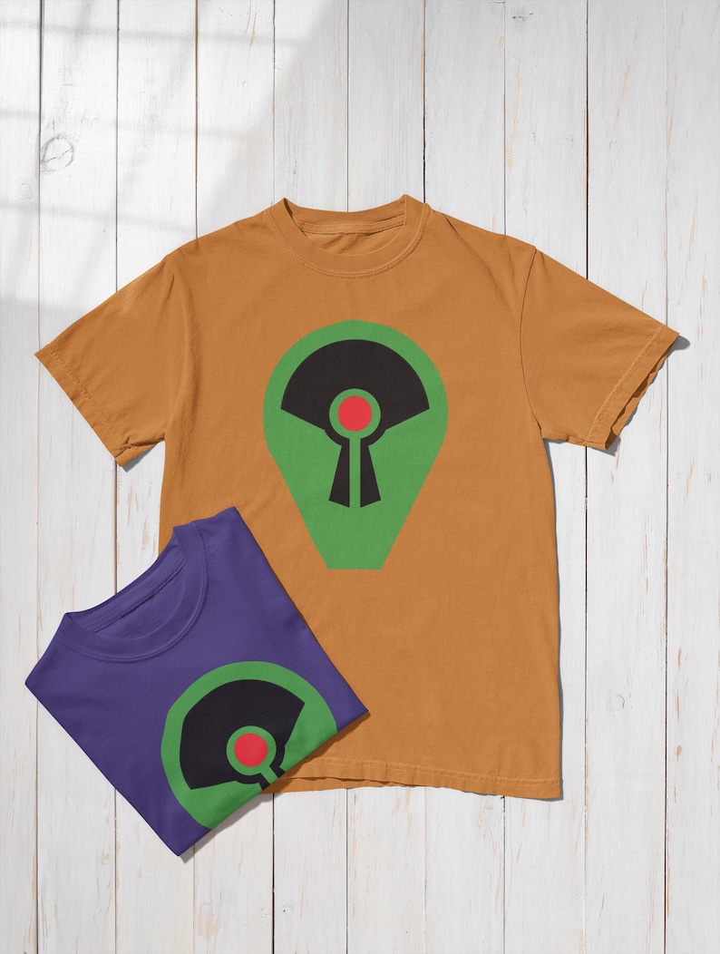 May include: Two t-shirts, one mustard yellow and one purple, are displayed on a white wooden surface. Each shirt features a graphic design with a green, black, and red abstract shape. The shirts are casual wear.