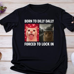 May include: A black t-shirt with the words "BORN TO DILLY DALLY FORCED TO LOCK IN". The shirt has two images of cats. One cat is sticking its tongue out, and the other is wearing a helmet.