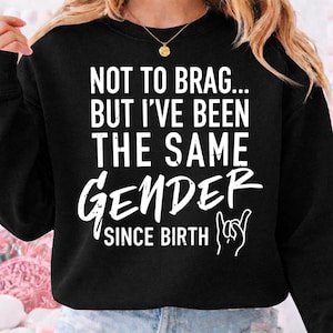 Op de afbeelding: Zwarte sweatshirt met witte tekst: "NOT TO BRAG... BUT I'VE BEEN THE SAME GENDER SINCE BIRTH" en een handgebaar. De top heeft een ronde hals en lange mouwen. Een gouden ketting is zichtbaar.