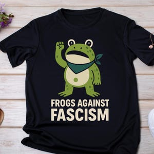 Puede incluir: Camiseta negra con una rana de dibujos animados con una bandana verde levantando el puño. La rana es verde con vientre color crema. El texto "FROGS AGAINST FASCISM" está impreso en blanco debajo. La camiseta es de material suave.
