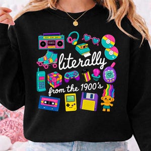 May include: A black long-sleeve sweatshirt featuring a colorful graphic print of 1990s nostalgia items including a boombox, roller skates, a Rubik's cube, a handheld game console, and a floppy disk with the text "literally from the 1900's".