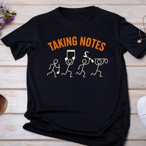 May include: Black t-shirt with the text "TAKING NOTES" in orange above a line of stick figures holding musical notes and symbols in cream. The shirt is made of a soft material.