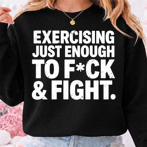 May include: Black sweatshirt with the white text: "EXERCISING JUST ENOUGH TO F*CK & FIGHT." The sweatshirt has a crew neck and long sleeves. A gold necklace with a pendant is visible.