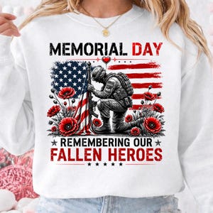 May include: White sweatshirt with a Memorial Day graphic. The design includes an American flag, a kneeling soldier, red poppies, and the text "MEMORIAL DAY REMEMBERING OUR FALLEN HEROES."