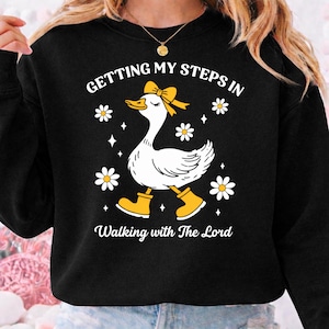 May include: Black sweatshirt with a whimsical graphic of a white goose wearing a yellow bow, boots, and surrounded by daisies. Text reads "Getting My Steps In" and "Walking with The Lord" in white script. A fun, casual design.