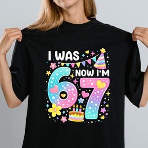 Może przedstawiać: Czarny t-shirt z kolorową grafiką na 67. urodziny. Projekt zawiera napis "I WAS...NOW I'M 67" z cyframi 6 i 7, sercami, kwiatami, czapeczką urodzinową i tortem.