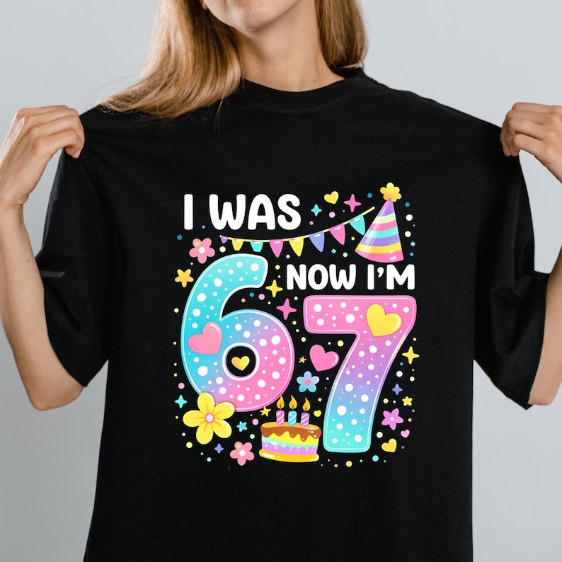 Może przedstawiać: Czarny t-shirt z kolorową grafiką na 67. urodziny. Projekt zawiera napis "I WAS...NOW I'M 67" z cyframi 6 i 7, sercami, kwiatami, czapeczką urodzinową i tortem.