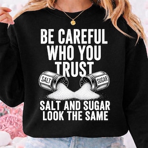 以下が含まれることがあります： 黒いスウェットシャツに、白い文字で「BE CAREFUL WHO YOU TRUST」と書かれています。その下に、塩と砂糖の容器が2つあり、1つには「SALT」、もう1つには「SUGAR」と書かれており、中身がこぼれています。「SALT AND SUGAR LOOK THE SAME」というテキストが下にあります。