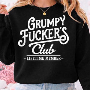 May include: Black sweatshirt with the text "Grumpy Fucker's Club - Lifetime Member" in white, stylized lettering. The sweatshirt is a solid black color, and the text is centered on the front.