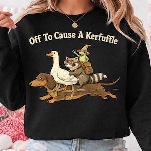Puede incluir: Sudadera negra con el texto "Off To Cause A Kerfuffle". El gráfico presenta un perro salchicha que lleva un ganso blanco, un mapache con gafas y una rana con sombrero de mago y mochila.