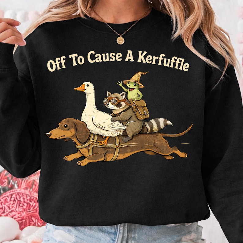 K&ouml;nnte beinhalten: Schwarzes Sweatshirt mit dem Text "Off To Cause A Kerfuffle". Die Grafik zeigt einen Dackel, der eine wei&szlig;e Gans, einen Waschb&auml;ren mit Brille und einen Frosch mit Hexenhut und Rucksack tr&auml;gt.