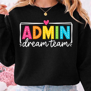 May include: A black crewneck sweatshirt featuring a graphic design with the word ADMIN in large, multicolored, polka-dotted letters, a small pink heart above, and the words dream team in white cursive script, all enclosed in a white rectangular frame.