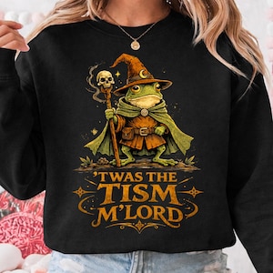 Op de afbeelding: Zwarte sweatshirt met een illustratie van een kikker-tovenaar met een staf en een hoed. De kikker draagt een cape en een riem. De tekst "'TWAS THE TISM M'LORD" staat eronder in oranje.