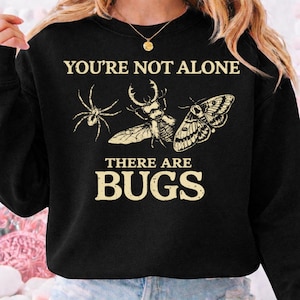 May include: Black sweatshirt with the text "YOU'RE NOT ALONE THERE ARE BUGS" in a cream color. The design features a spider, beetle, and moth. The sweatshirt is a crew neck style.
