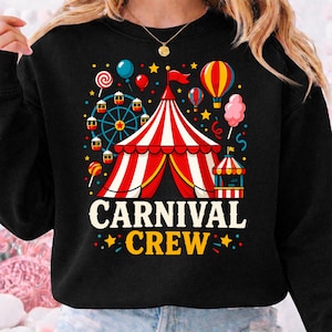 Pode incluir: Camisola preta com um gráfico de carnaval colorido. O design apresenta uma tenda listrada vermelha e branca, uma roda gigante, balões e as palavras "CARNIVAL CREW" em amarelo e branco.
