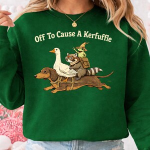 K&ouml;nnte beinhalten: Dunkelgr&uuml;nes Sweatshirt mit dem wei&szlig;en Schriftzug "Off To Cause A Kerfuffle". Die Grafik zeigt einen Dackel, der eine wei&szlig;e Gans, einen Waschb&auml;ren mit Brille und einen Frosch mit Hexenhut tr&auml;gt.