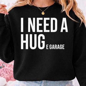 May include: A black long-sleeve crewneck sweatshirt featuring the white text I NEED A HUGE GARAGE printed on the front. The garment has a ribbed neckline and cuffs, and is worn with a gold-toned chain necklace with a small circular pendant.
