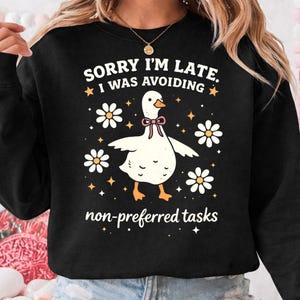 May include: Black sweatshirt with a white goose graphic wearing a pink bow tie, surrounded by white daisies and stars. Text reads "SORRY I'M LATE, I WAS AVOIDING non-preferred tasks."