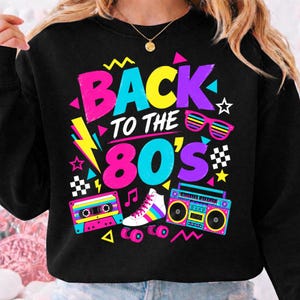 Pode incluir: Uma camisola preta com um gráfico vibrante e o texto BACK TO THE 80'S em rosa, amarelo e azul néon. O design inclui uma cassete, um patim, um rádio leitor de cassetes e óculos de sol, acentuados por formas geométricas e estrelas coloridas.