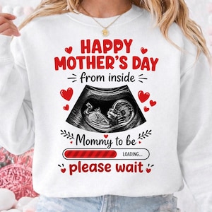 Può includere: Felpa bianca con una grafica che celebra la festa della mamma. Il design include cuori rossi, il testo "Happy Mother's Day from inside" e un'immagine ecografica di un feto. Sono incluse anche le frasi "Mommy to be" e "please wait".