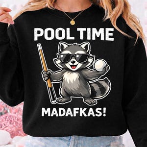 May include: Black sweatshirt with the text "POOL TIME MADAFKAS!". The graphic features a raccoon wearing sunglasses, holding a pool cue and a white billiard ball. The raccoon is gray, white, and black.