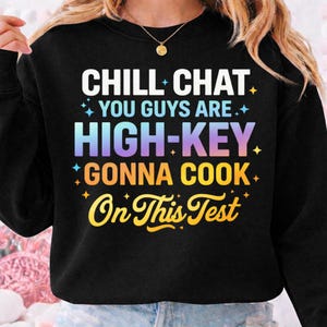 May include: Black sweatshirt with the text "CHILL CHAT YOU GUYS ARE HIGH-KEY GONNA COOK On This Test" in white, blue, yellow, and orange. The text has a gradient effect. The sweatshirt is a crew neck.