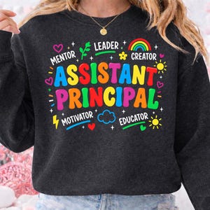 Może przedstawiać: Ciemnoszara bluza z napisem "ASSISTANT PRINCIPAL" dużymi, kolorowymi literami. Projekt zawiera słowa "MENTOR", "LEADER", "CREATOR", "MOTIVATOR" i "EDUCATOR", a także serca, tęcze i słońca.