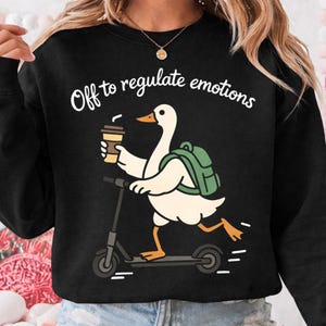 May include: Black sweatshirt with the text "Off to regulate emotions." The graphic shows a white goose riding a scooter, carrying a coffee cup and a green backpack. The goose has orange feet and a yellow beak. The design is a fun, whimsical illustration.