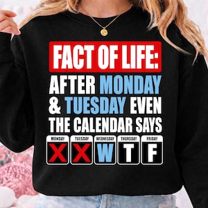 Puede incluir: Una sudadera negra de manga larga con el texto "FACT OF LIFE: AFTER MONDAY & TUESDAY EVEN THE CALENDAR SAYS" en blanco y azul claro. Debajo, cinco cuadrados de calendario muestran "MONDAY" y "TUESDAY" con marcas X rojas, seguidos de "W T F".