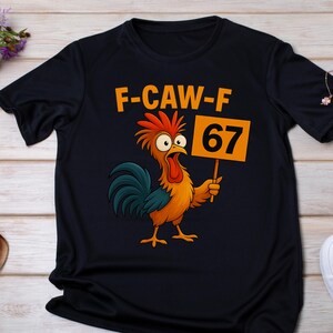 May include: Black t-shirt featuring a cartoon rooster with orange, yellow, and blue feathers, holding a sign that reads "67". Above the rooster, the text "F-CAW-F" is printed in orange. The shirt is made of a soft material.