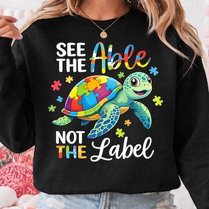 May include: Black sweatshirt with a colorful turtle design. The turtle's shell is a jigsaw puzzle, and the text reads "SEE THE Able NOT THE Label". Colorful puzzle pieces and stars surround the turtle.