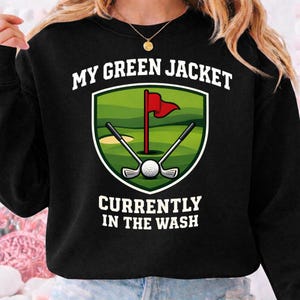 May include: Black sweatshirt with a golf-themed graphic. The graphic features a green shield with a golf course scene, a red flag, golf clubs, and a golf ball. Text reads "MY GREEN JACKET CURRENTLY IN THE WASH."