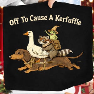 K&ouml;nnte beinhalten: Schwarzes T-Shirt mit dem Text "Off To Cause A Kerfuffle". Die Illustration zeigt einen Dackel, eine Gans, einen Waschb&auml;ren mit Brille und einen Frosch mit Hexenhut.